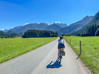 Radfahrer auf asphaltiertem Weg durch grüne Wiesen mit Alpenbergen im Hintergrund an sonnigem Tag am Salzachradweg bei Kuchl.