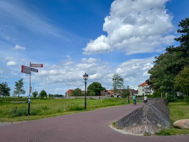 Red brick cycling path in Plantagepark, Harderwijk, with cyclists, directional signs, street lamp, and green parkland under blue sky with clouds.
