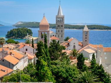 Old town of Rab with four distinctive bell towers rising above terracotta roofs, surrounded by lush greenery and the Adriatic Sea.