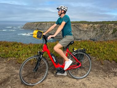 Radfahrer auf rotem E-Bike mit gelber Tasche fährt entlang der Küstenklippen bei Cavaleiro. Felsige Klippen und Meer im Hintergrund.