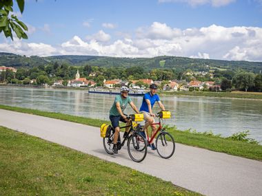 Two cyclists ride along the Danube, with the village of Persenbeug-Gottsdorf in the background