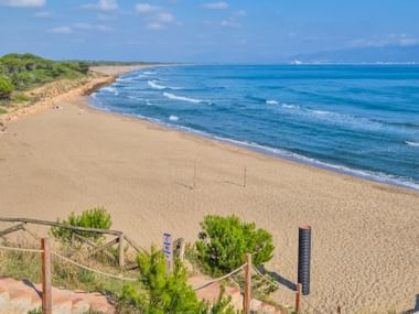 Breiter Sandstrand in Katalonien mit türkisfarbenem Mittelmeer, sanften Wellen und grüner Vegetation entlang der Küste unter blauem Himmel.