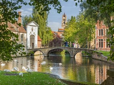 Steinbrücke über Kanal in Brügge mit Schwänen, historischen Gebäuden, weißer Kapelle und Kirchturm. Bäume rahmen die Szene unter blauem Himmel.