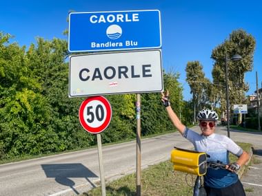 Radfahrer mit Helm und Fahrrad zeigt auf Ortsschild von Caorle mit Bandiera Blu Symbol. Geschwindigkeitsbegrenzung 50 darunter.