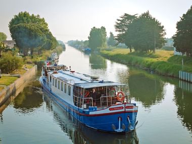 Blau-weißes Passagierschiff MS Lestello am Ufer eines von Bäumen gesäumten Kanals mit grünen Ufern. Menschen auf Deck unter bewölktem Himmel.