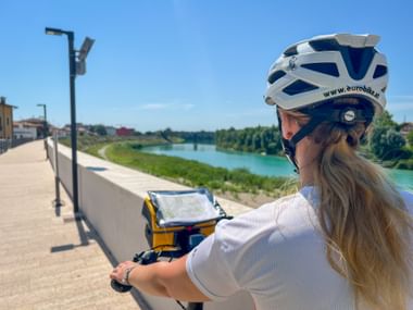 Radfahrerin mit weißem Helm prüft Karte am Lenker entlang des Latisana Radwegs neben türkisfarbenem Fluss unter blauem Himmel in Friaul.
