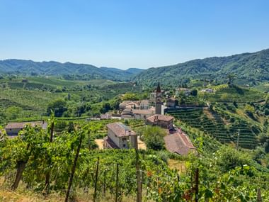 Italienisches Dorf mit Kirchturm inmitten terrassierter Prosecco-Weinberge bei Conegliano, mit grünen Hügeln unter blauem Himmel.
