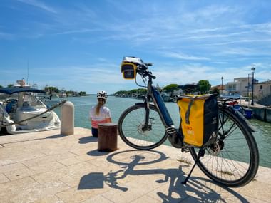 Fahrrad mit gelben Taschen am Hafen von Marano Lagunare. Radfahrer sitzt auf Poller und blickt auf Boote und Uferpromenade bei blauem Himmel.