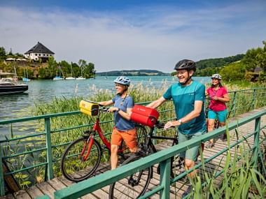 Three cyclists with helmets crossing a green wooden pier at Mattsee lake. Sailboats and reeds visible in the turquoise water, hills in background.