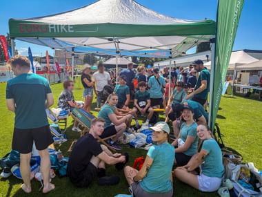 Team members in turquoise shirts gathered under a green Eurohike tent at an outdoor sports event, sitting and standing on grass under blue sky.