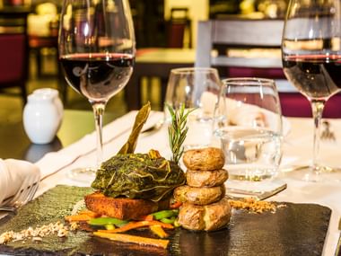 Elegantes portugiesisches Bacalhau da Couvada auf schwarzem Schiefer mit Röstkartoffeln, Gemüse und Kräutern, begleitet von Rotweingläsern.
