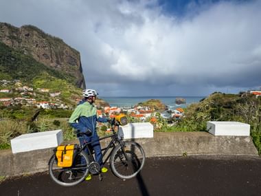 Cyclist with yellow panniers at coastal viewpoint overlooking Porto da Cruz village, dramatic cliff, and Atlantic Ocean under cloudy sky.
