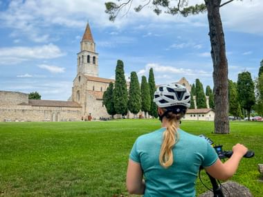 Radfahrerin mit weißem Helm und türkisfarbenem Shirt blickt auf die Basilika Santa Maria Assunta in Aquileia über grüne Wiese mit Zypressen.