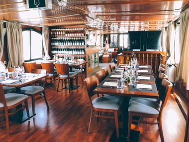 Innenraum des MS Lestello Salons mit Holztischen, gepolsterten Stühlen, Weinregal an der Wand und großen Fenstern mit Vorhängen.