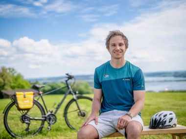 Nicolai Warren im blauen Radtrikot sitzt auf Holzbank mit Fahrrad und Helm neben sich, mit Blick auf grüne Landschaft und See.