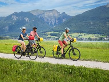 Three cyclists with panniers riding on a lakeside path near Wolfgangsee in Salzkammergut, with mountains and a village in the background.