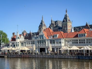 Uferrestaurants mit weißen Sonnenschirmen entlang der Prins Hendrikkade in Amsterdam mit traditionellen niederländischen Gebäuden.