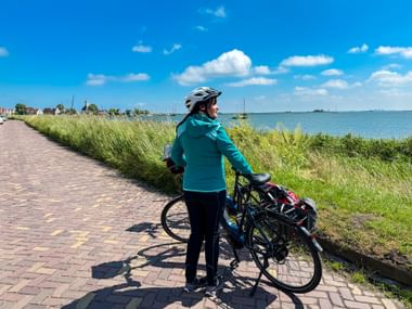 Radfahrerin in türkisfarbener Jacke und Helm steht mit Fahrrad auf Ziegelweg am IJsselmeer, mit Booten und blauem Himmel sichtbar.