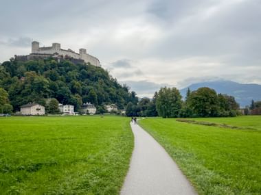 Paved cycling path through green meadows leading towards Hohensalzburg Fortress on a hilltop, with mountains in the background under cloudy skies.