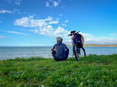 Cyclist sitting on grass beside touring bike, overlooking the blue sea at Baratti bay. Green grass in foreground, calm water and coastline visible.