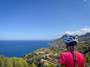 Radfahrerin in rosa Shirt und Helm betrachtet Mittelmeerküste von erhöhtem Aussichtspunkt in Banyalbufar, Mallorca.