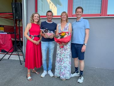 Vier Personen stehen zusammen im Freien und halten Geschenkkörbe und Blumen. Zwei Frauen in Kleidern flankieren zwei Männer in Freizeitkleidung.