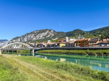 Arched pedestrian bridge crossing turquoise river in San Michele all'Adige with colorful buildings, church tower, and mountains in background.