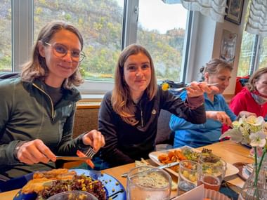 Vier Personen genießen Mittagessen am Restauranttisch mit Tellern voller Essen und Getränken, große Fenster zeigen Herbstlandschaft.