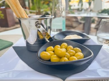 Dunkle Schale mit grünen Oliven, Olivenölflasche im Eiskübel und Grissini auf einem Restauranttisch in Port d'Andratx, Mallorca.