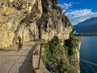 Cyclist in yellow jersey riding along Strada del Ponale, a scenic cliff path carved into rock face above Lake Garda with mountains in background.