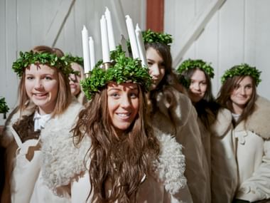 Gruppe von Mädchen in weißen Gewändern mit grünen Kränzen. Das vordere Mädchen trägt eine Krone mit sieben weißen Kerzen und lächelt.