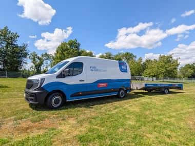 White and blue Eurobike e-bus with bicycle trailer parked on grass field under blue sky with trees in background.