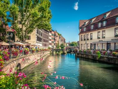 Canal in La Petite France quarter, Strasbourg, with colorful half-timbered houses, pink flowers, and people at waterside cafes under blue sky.