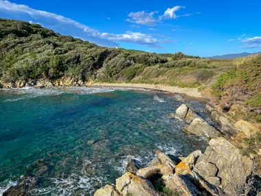Scenic bay Baia del Pozzino near Baratti with turquoise water, rocky coastline, pebble beach, and green hills under blue sky.