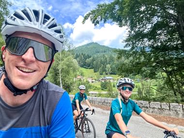 Thomas and family taking a selfie on the Alpe Adria cycle route near Tarvis, with forested mountains and alpine village in the background.