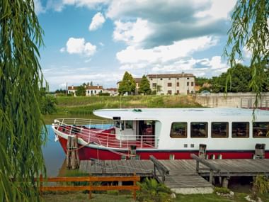 MS Ave Maria, a red and white passenger ship, moored at a wooden dock in Portegrandi. Willow trees frame the scene with village buildings visible.
