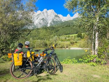 Two cyclists with yellow panniers rest by a green lake near Pfarrwerfen. Forested mountains rise behind them under a blue sky.