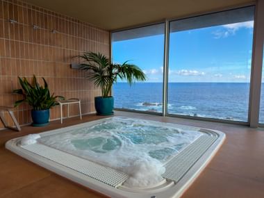 Indoor whirlpool with bubbling water and large windows overlooking the Atlantic Ocean in Porto Moniz, Madeira. Two potted plants decorate the tiled room.