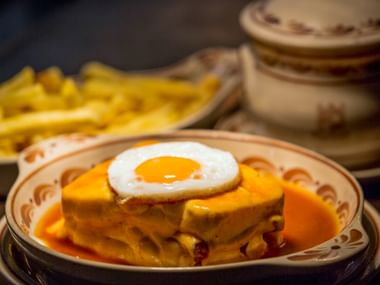 Traditionelles portugiesisches Francesinha-Sandwich mit Spiegelei und oranger Käsesauce, serviert auf dekorativem Teller mit Pommes im Hintergrund.