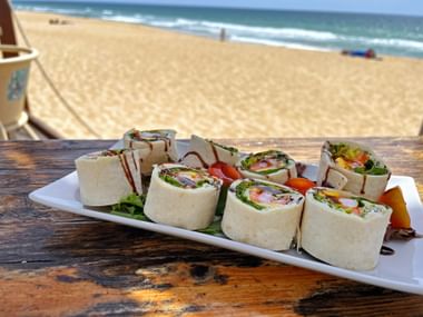 Teller mit geschnittenen Wrap-Rollen mit Gemüse auf Holztisch am Strand. Sandstrand und Meereswellen im Hintergrund sichtbar.