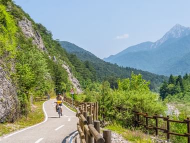 Cyclist on paved bike path with wooden fence in Kanaltal valley. Green mountains and rocky cliffs visible under blue sky.