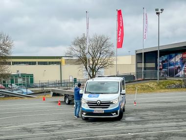 White Renault van on training course with orange cones. Person in blue jacket stands beside vehicle on marked parking lot. Eurofun flags visible.