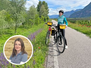 Woman with bicycle on paved path through green meadows with mountains in background. Inset portrait shows smiling woman in gray polo shirt.