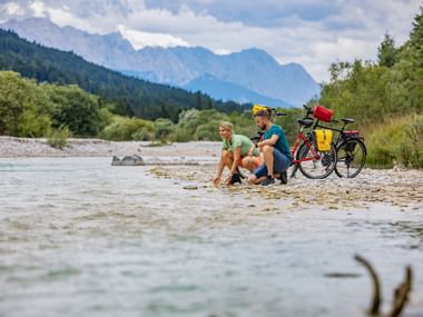 Two cyclists sitting on rocky riverbank of the Lech River with their touring bicycles, surrounded by alpine mountains and green forests.