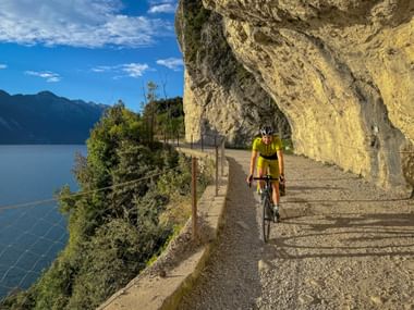 Cyclist in yellow jersey riding along Strada del Ponale, a scenic path carved into cliff above Lake Garda with mountains in background.