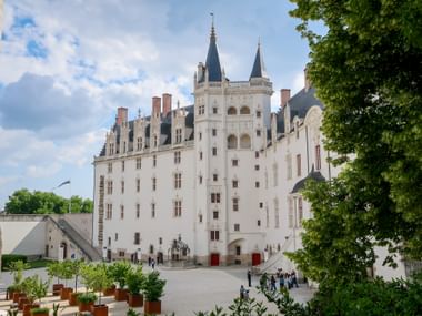 White castle with twin spired towers and courtyard in Nantes. Visitors walk across the square framed by green trees and potted plants.