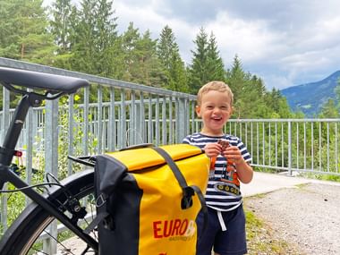 Young boy in striped shirt eating a cookie on a bridge along the Drau cycle path. Yellow Eurobike pannier bag on bicycle, forest and mountains visible.