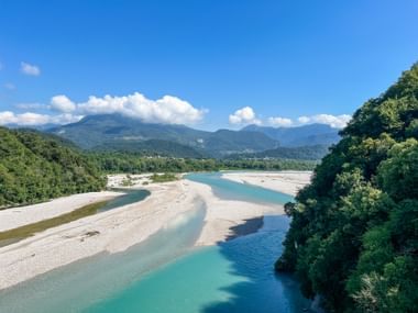 Tagliamento-Flussbett mit türkisfarbenen Wasserläufen durch weiße Kiesbänke, umgeben von grünen Wäldern und Bergen unter blauem Himmel.