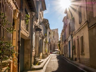Schmale Gasse in Arles mit historischen Steingebäuden auf beiden Seiten, hellem Sonnenlicht und einer Kirchenruine am Ende der Straße.