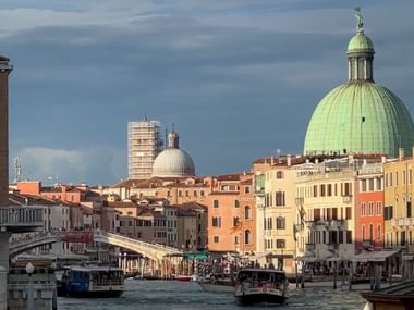View of Venice's Canal Grande with boats, colorful buildings, and a prominent green-domed church under a cloudy sky.